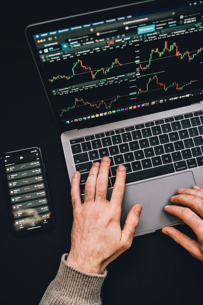 Mains en train de taper sur un ordinateur portable, avec des graphiques de cryptomonnaies et l’écran d’un smartphone visibles, en mettant l’accent sur la finance numérique.